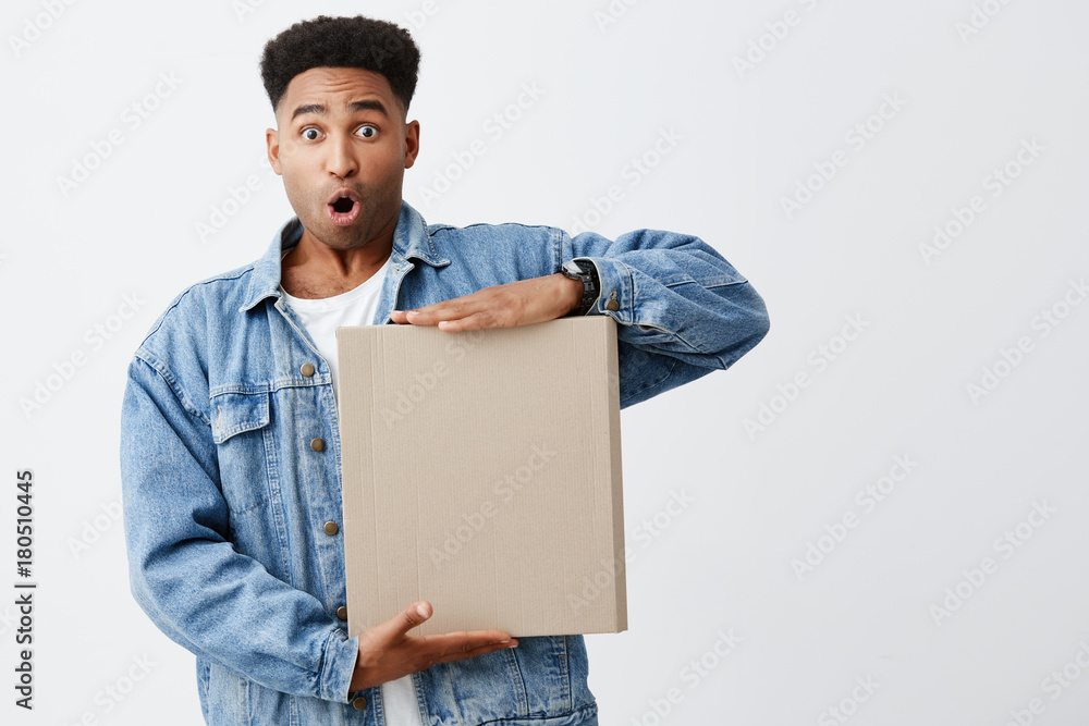 What is it. Isolated on white portrait of beautiful dark-skinned funny male student with afro hairstyle in white t-shirt and denim jacket holding box in hands with surprise and curious expression What is it. Isolated on white portrait of beautiful dark-skinned funny male student with afro hairstyle in white t-shirt and denim jacket holding box in hands with surprise and curious expression