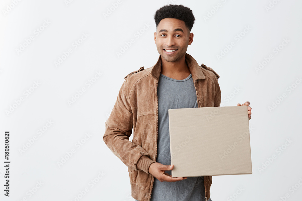 Positive emotions. Close up of young beautiful black-skinned male with afro hairstyle in casual stylish clothes holding cardboard in hands, looking in camera with happy and relaxed expression. Positive emotions. Close up of young beautiful black-skinned male with afro hairstyle in casual stylish clothes holding cardboard in hands, looking in camera with happy and relaxed expression.