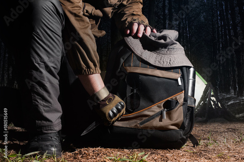 Canvastavla Man in tactical outfit holding a knife and kneeling for backpack with camping and tactical gear on night forest background