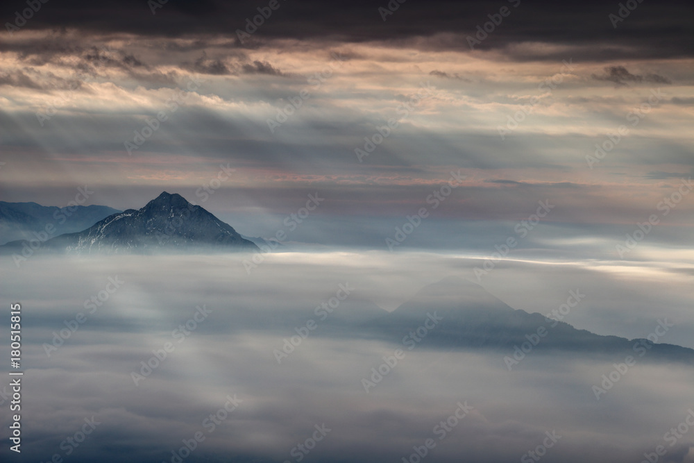 Curtain of sun rays shining through dark gray clouds illuminates two conical mountains in autumn morning mist, Storzic and Tolsti vrh peaks, Kamnik Savinja Alps, Gorenjska, Carniola, Slovenia, Europe 