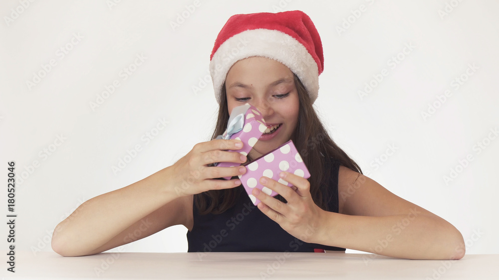 Beautiful teen girl in Santa Claus hat sitting and joyfully opens the gift box on white background Beautiful teen girl in Santa Claus hat sitting and joyfully opens the gift box on white background