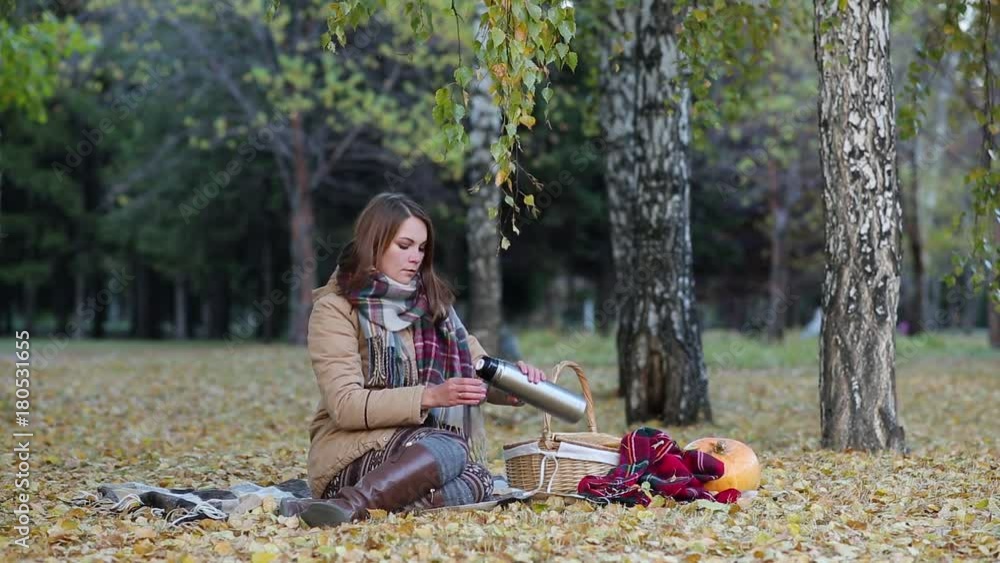 young woman drinking tea from a thermos in the autumn Park sitting on a blanket