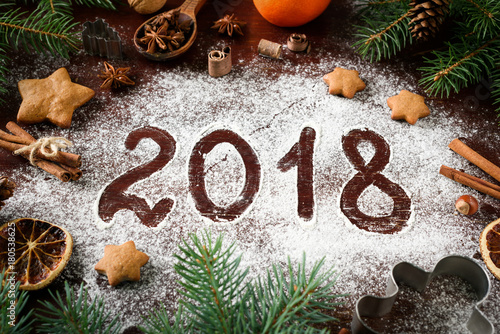 New Year 2018 written on flour and Christmas Decorations Gingerbread cookies, cinnamin, oranges, spices, nuts and cookie cutters on wooden background. Christmas card, New Year greeting card