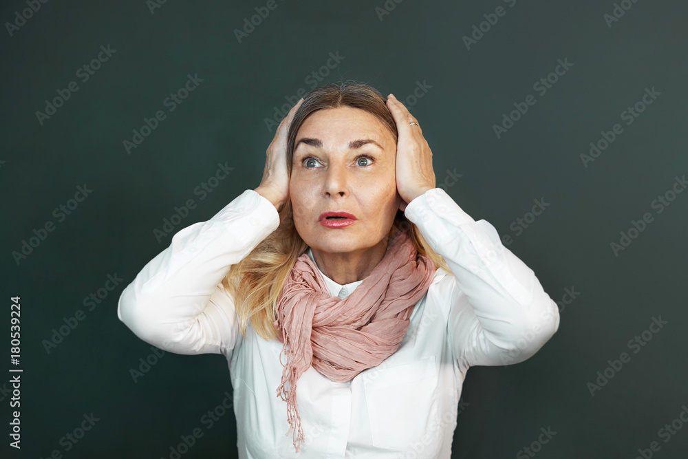 Isolated shot of emotional mature 60 year old Caucasian female wearing white blouse and pink scarf having frustrated look, covering ears with both hands, can't stand unpleasant sound or noise Isolated shot of emotional mature 60 year old Caucasian female wearing white blouse and pink scarf having frustrated look, covering ears with both hands, can't stand unpleasant sound or noise