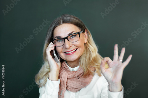 Attractive confident mature businesswoman with loose blonde hair and stylish scarf wrapped around her neck smiling, making ok gesture, having successful business negotiations using mobile phone