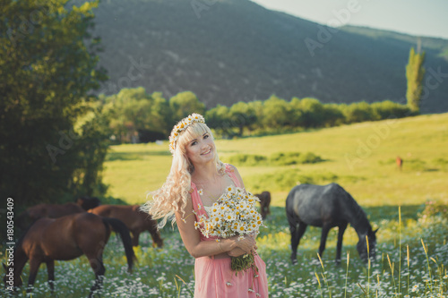 Seductive blond blue eyes lady woman in pinky airy dress on meadow of daisy chamomile holding bouquet of nflowers in hands and posing with horse on background.Adorable scene in nature. photo