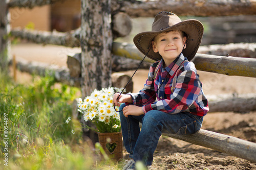 Cute fermer handsome boy cowboy in jeans enjoying summer day in village life with flowers wearing leather cow hat happyly smiling.