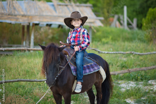 Cute fermer handsome boy cowboy in jeans enjoying summer day in village life with flowers wearing leather cow hat happyly smiling and sitting on horse .