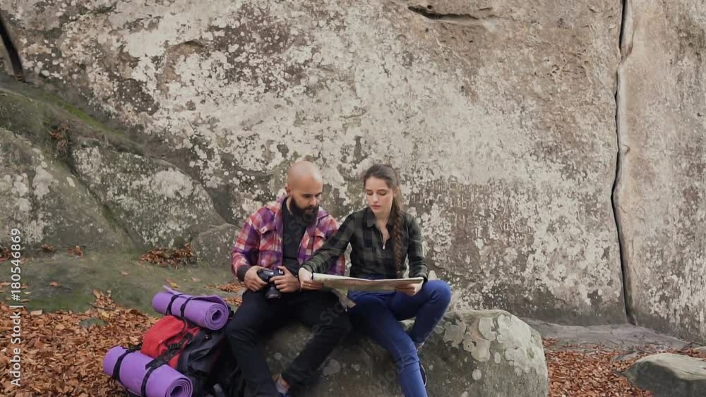 Young happy couple of tourists are studying a map, sitting on a stone near a big rock, a man and a woman backpackers, enjoying leisure while hiking in the mountains Young happy couple of tourists are studying a map, sitting on a stone near a big rock, a man and a woman backpackers, enjoying leisure while hiking in the mountains