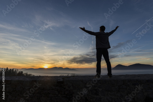 Man with his arms spread widely standing outdoors silhouetted against a colorful sunrise