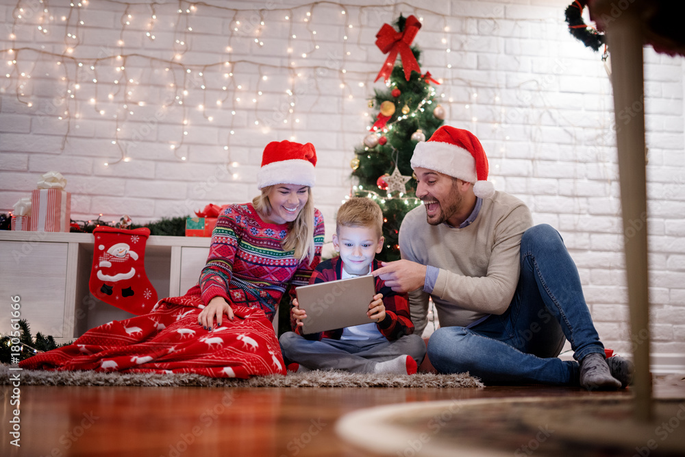 Fototapeta premium Positive young beautiful parents spending a great time with their happy kid while sitting on a carpet and looking at a tablet in the warm house for Christmas holidays.