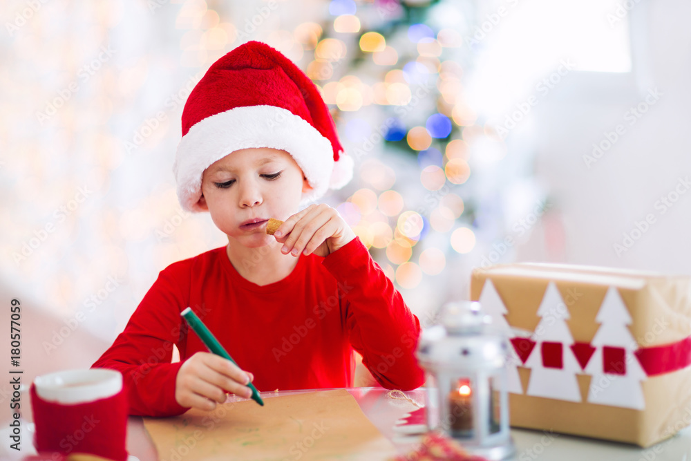 Boy writing letter to Santa Claus in red hat near the Christmas tree Boy writing letter to Santa Claus in red hat near the Christmas tree
