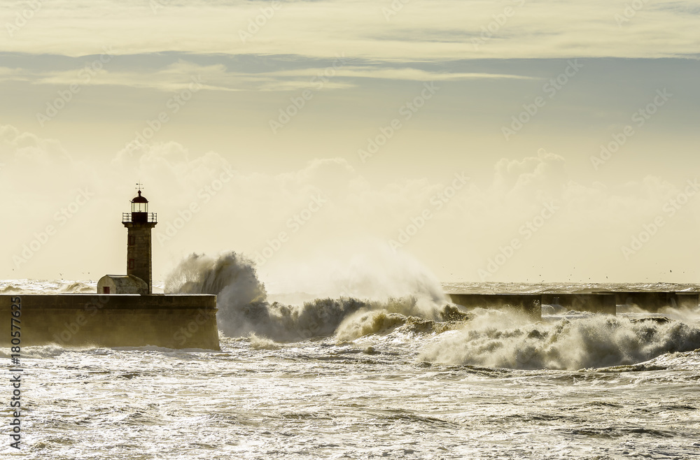 Landscape seascape lighthouse battered by huge waves on Atlantic Ocean with blue green skies and puffy clouds.