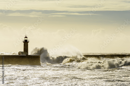 Landscape seascape lighthouse battered by huge waves on Atlantic Ocean with blue green skies and puffy clouds.