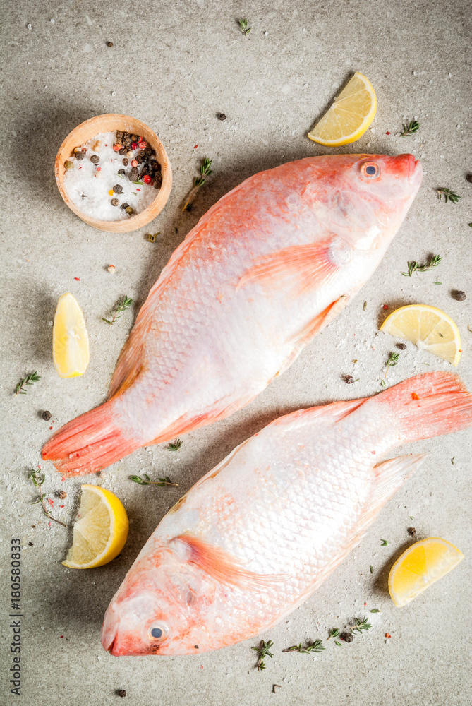 Fresh raw fish pink tilapia with spices for cooking - lemon, salt, pepper, herbs, on gray stone table, copy space