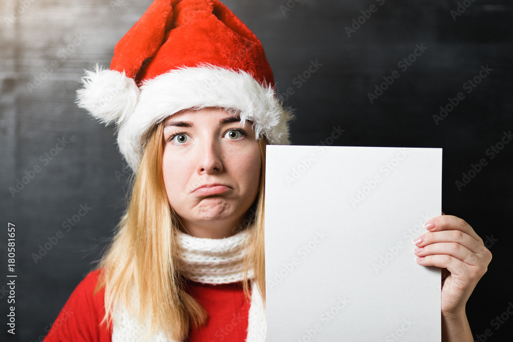 Portrait of a beautiful sad girl in Santa Claus hat red shirt and knitted scarf holding a white sheet of paper with place for text Portrait of a beautiful sad girl in Santa Claus hat red shirt and knitted scarf holding a white sheet of paper with place for text