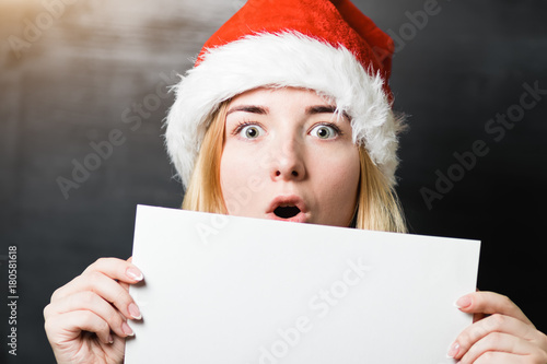 Portrait of a beautiful shocked girl in Santa Claus hat red shirt and knitted scarf holding a white sheet of paper with place for text © Romvy