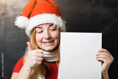 Portrait of a beautiful cheerful girl in Santa Claus hat red shirt and knitted scarf holding a white sheet of paper with place for text © Romvy