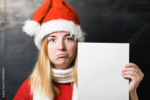 Portrait of a beautiful sad girl in Santa Claus hat red shirt and knitted scarf holding a white sheet of paper with place for text © Romvy