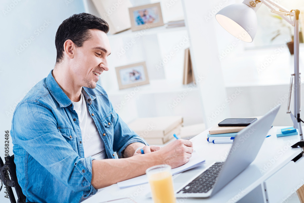 Making notes. Joyful young man keeping smile on his face and bowing head while sitting in semi position Making notes. Joyful young man keeping smile on his face and bowing head while sitting in semi position