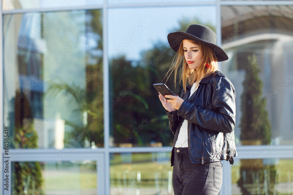 Stylish hipster girl wears black leather jacket and holding phone before modern building
