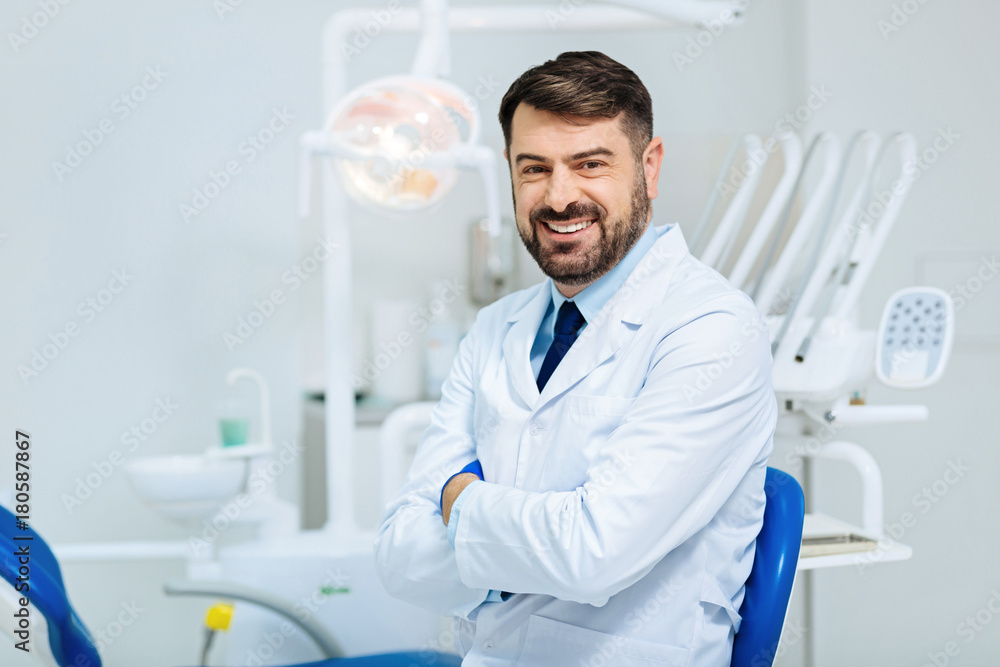 Healthy smile. Positive bearded dentist expressing cheer while standing in front of you and keeping arms in crossed position Healthy smile. Positive bearded dentist expressing cheer while standing in front of you and keeping arms in crossed position
