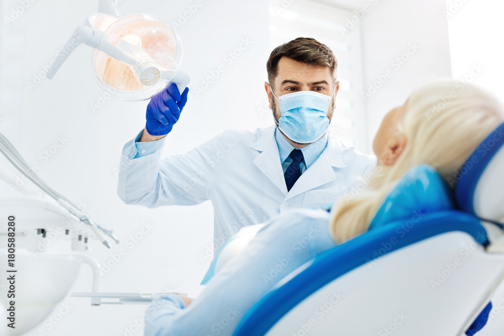Do not worry. Optimistic dentist with a face mask standing near the patient while touching medical tool Do not worry. Optimistic dentist with a face mask standing near the patient while touching medical tool
