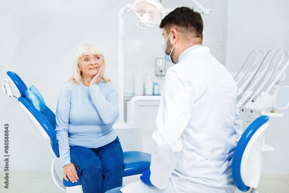Obraz premium Feeling discomfort. Sad woman having medical consultation while sitting on the dental chair and touching her cheek
