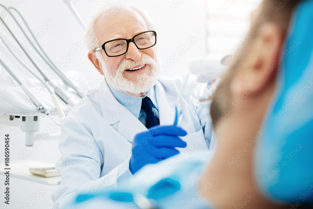 Working hours. Close up of experienced dentist sitting near the patient while holding false teeth with tooth brush and expressing cheer Working hours. Close up of experienced dentist sitting near the patient while holding false teeth with tooth brush and expressing cheer