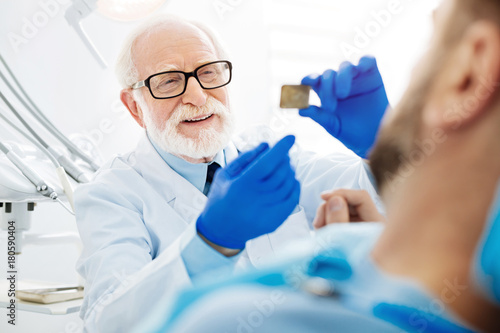 Working with pleasure. Close up of professional dentist holding Xray image in hand and being in cheerful mood while the patient lying on the couch