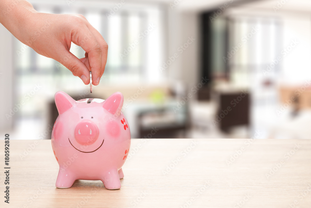 Hand putting a coin into a pink piggy bank on wooden desk on home background - save money concept. Hand putting a coin into a pink piggy bank on wooden desk on home background - save money concept.