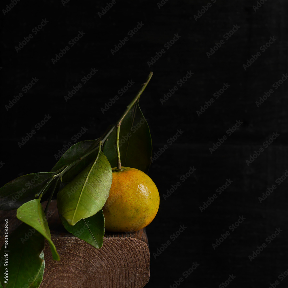 One mandarin on a branch with green leaves on a dark background with copy space. Citrus fruit in a low key on a wooden board. Black minimal background. Composition with tangerine. One mandarin on a branch with green leaves on a dark background with copy space. Citrus fruit in a low key on a wooden board. Black minimal background. Composition with tangerine.