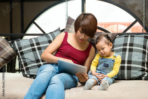 Mother showing media content on line to her twenty months daughter in a tablet sitting on a couch in the living room in a house interior © supong32