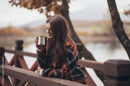 Young beautiful woman in a warm classic coat drinking tea near lake. Autumn atmosphere