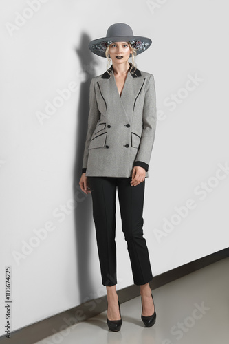 Pretty fashion model in jacket, pants and broad-brim hat with flowers posing near gray wall