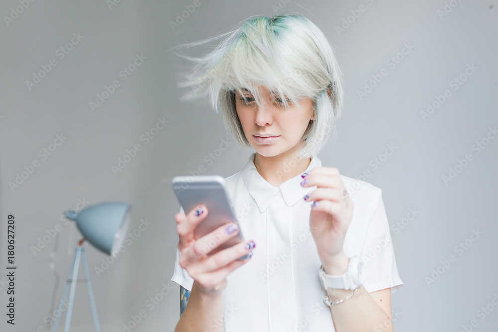 Portrait of cute girl with gray short hairstyle standing on gray background. She wears white dress and light makeup, phone in hands. Her hair is flying from wind. Portrait of cute girl with gray short hairstyle standing on gray background. She wears white dress and light makeup, phone in hands. Her hair is flying from wind.