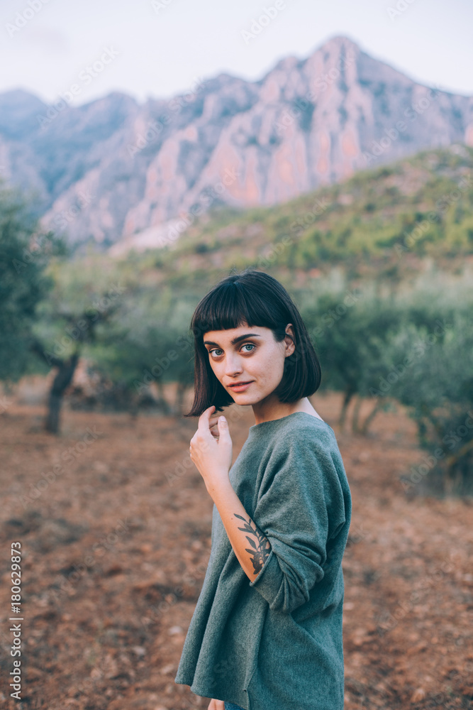 Portrait of adorable and attractive woman or young traveling female smiling to camera, tranquil and calm while exploring national park forest with mountains and olive trees, concept hipster fashion Portrait of adorable and attractive woman or young traveling female smiling to camera, tranquil and calm while exploring national park forest with mountains and olive trees, concept hipster fashion