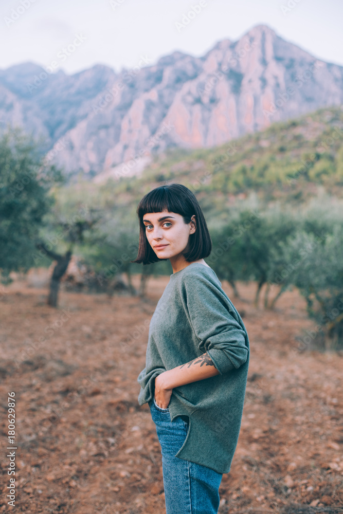 Naklejka premium Young female tourist, pretty beautiful woman with brown hair, with travel backpack wears comfortable wool sweater, smiles to camera with mountains and trees in background
