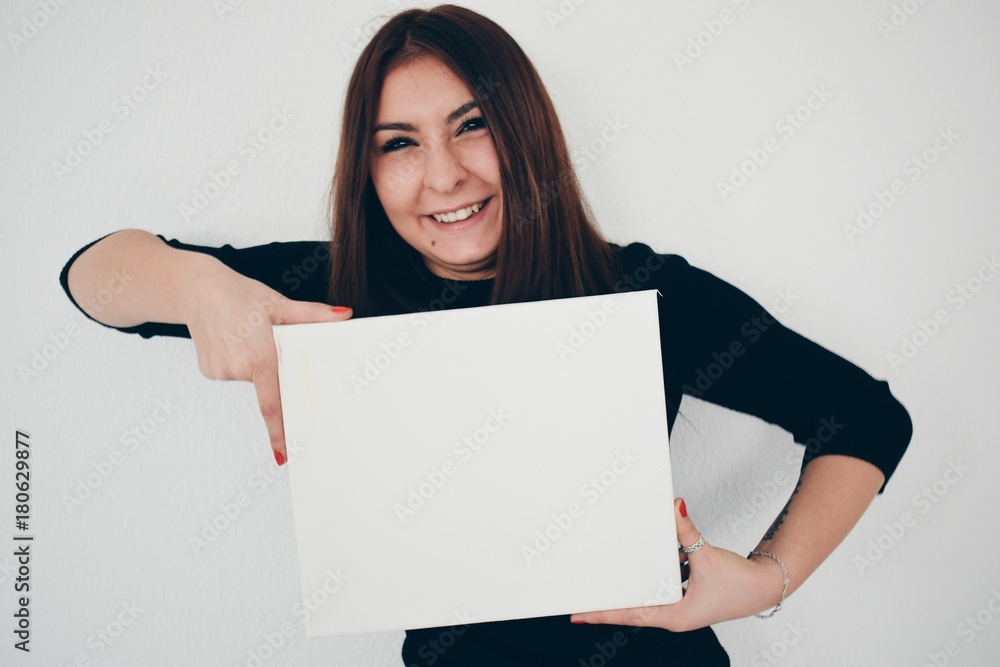 on light background is standing young, smiling woman, holding empty white board and pointing at with her finger on light background is standing young, smiling woman, holding empty white board and pointing at with her finger