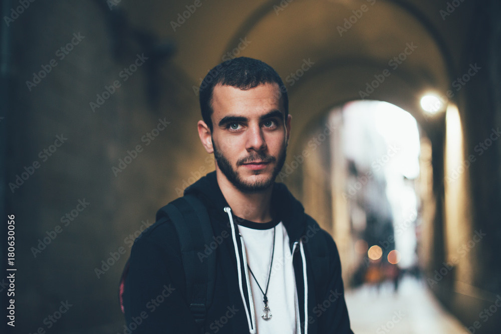 Obraz premium Handsome attractive young man in 20s, student or professional freelancer, looks into camera, stands in dark street or alley in Barcelona, exchange education program, confident and calm