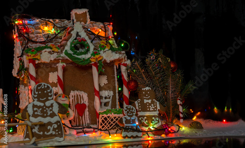 Gingerbread family near snow-covered homemade gingerbread house with Christmas lights on dark background