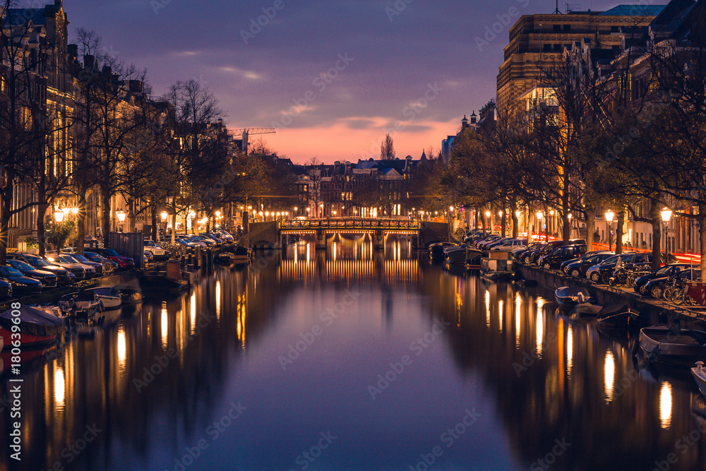 Purple cloudy sky, reflections in the water, boats and cosy-lighted streets during a twilight blue hour sunset, Amsterdam, Netherlands