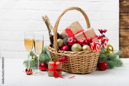 Two glasses of champagne with Christmas basket with bottle, gifts with red satin ribbon, candy canes, pine cones, golden garlands on white brick wall background, new year celebration concept