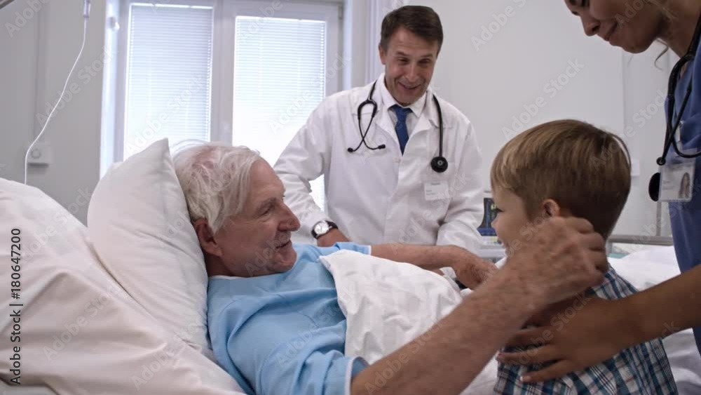 Senior man lying on comfortable bed in private hospital ward, talking with doctor and then hugging little grandson while nurse taking care of him Senior man lying on comfortable bed in private hospital ward, talking with doctor and then hugging little grandson while nurse taking care of him