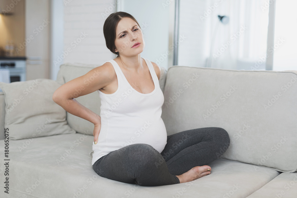 A pregnant woman sits at home on a light sofa. Her back hurts and she holds on to her A pregnant woman sits at home on a light sofa. Her back hurts and she holds on to her