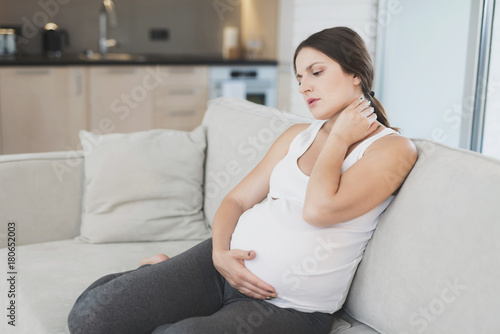 A pregnant woman sits at home on a light sofa. She has a stiff neck and she rubs it. Free Download A pregnant woman sits at home on a light sofa. She has a stiff neck and she rubs it. - GFXLoad