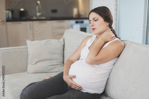 A pregnant woman sits at home on a light sofa. She has a stiff neck and she rubs it. Free Download A pregnant woman sits at home on a light sofa. She has a stiff neck and she rubs it. - GFXLoad