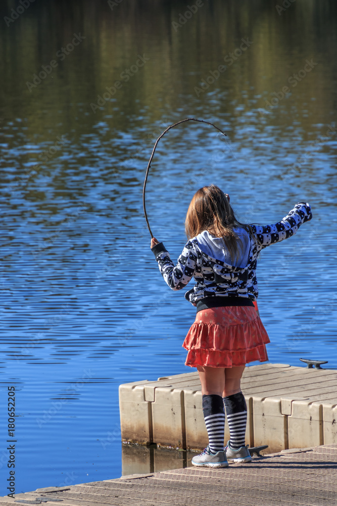 A little girl fishing from a dock on a lake. She has red shorts, black long  socks and blue and white jacket. She is fishing with a stick. Stock Photo