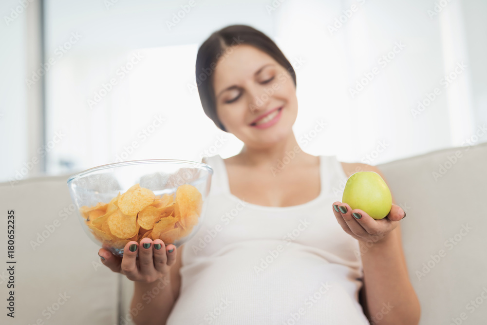 A pregnant woman is sitting on a light sofa at home. She holds a vase of chips and an apple in her hands A pregnant woman is sitting on a light sofa at home. She holds a vase of chips and an apple in her hands