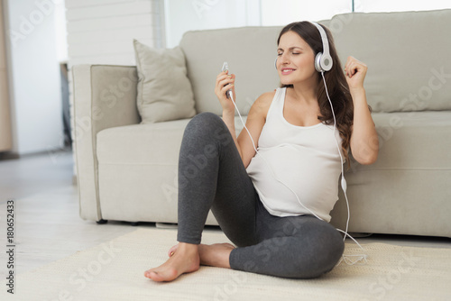 A pregnant woman sits on a light floor at home. She wore headphones and listened to music